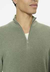 Pull vert à fermeture éclair avec tissu tricoté texturé. Doté d'un col montant et d'une fermeture éclair en métal. Sous-vêtement visible de couleur blanc cassé.