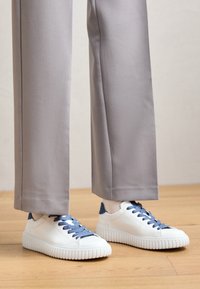 Bílé tenisky s modrými tkaničkami a modrými detaily na patě, s texturovanou gumovou podrážkou, doplněné světle šedými kalhotami s širokými nohavicemi.