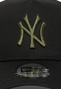 Melna cepure ar izšūtu olīvu zaļu "NY" logotipu priekšpusē. Regulējama siksna un New Era uzlīme uz cepures apakšpuses.