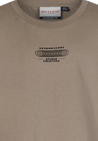 Beige Baumwoll-T-Shirt mit kurzen Ärmeln, das ein schwarzes grafikdesign zeigt, das die Texte "RETOUR JEANS" und "STUDIO CREATIVES" enthält.