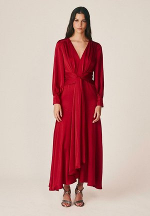 Femme debout portant une longue robe rouge à manches longues avec un décolleté en V profond, nouée à la taille, associée à des sandales à talons hauts à brides.