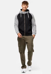 Sudadera con capucha y cremallera de cuerpo negro y mangas grises, combinada con pantalones cargo en color oliva. Cuenta con cordones y cierre frontal de cremallera.