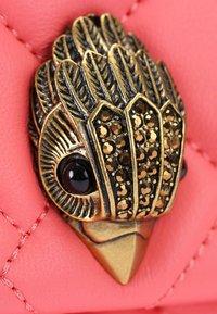 Accento di uccello in bronzo con design dettagliato delle piume e gemme nere, montato su uno sfondo rosa trapuntato, creando un contrasto testurizzato.