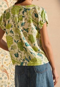 Blomstret bluse med en cremefarvet baggrund og grønne samt blå botaniske mønstre. Korte pufærmer med en knaplukning bagpå.