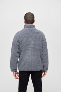 Hallhall mustri ja kõrge kaelusega hall fliisist pullover, millel on tekstureeritud pind ja pikad varrukad. Kantud mustade pükste peal, minimalistlik disain ilma nähtavate logodeta.
