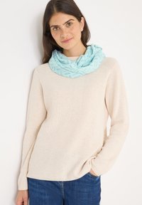 Lichtblauwe, gemusterde infinitiesjaal van zachte stof, gedragen over een beige geribde trui, gecombineerd met blauwe jeans.