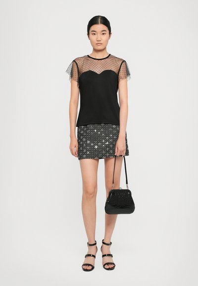 Sieviete tērpta melnā caurspīdīgā polka punktu blūzē, melnā sequin mini svārkā, melnās strappy kurpes, tur rokās mazu melnu somiņu uz balta fona.