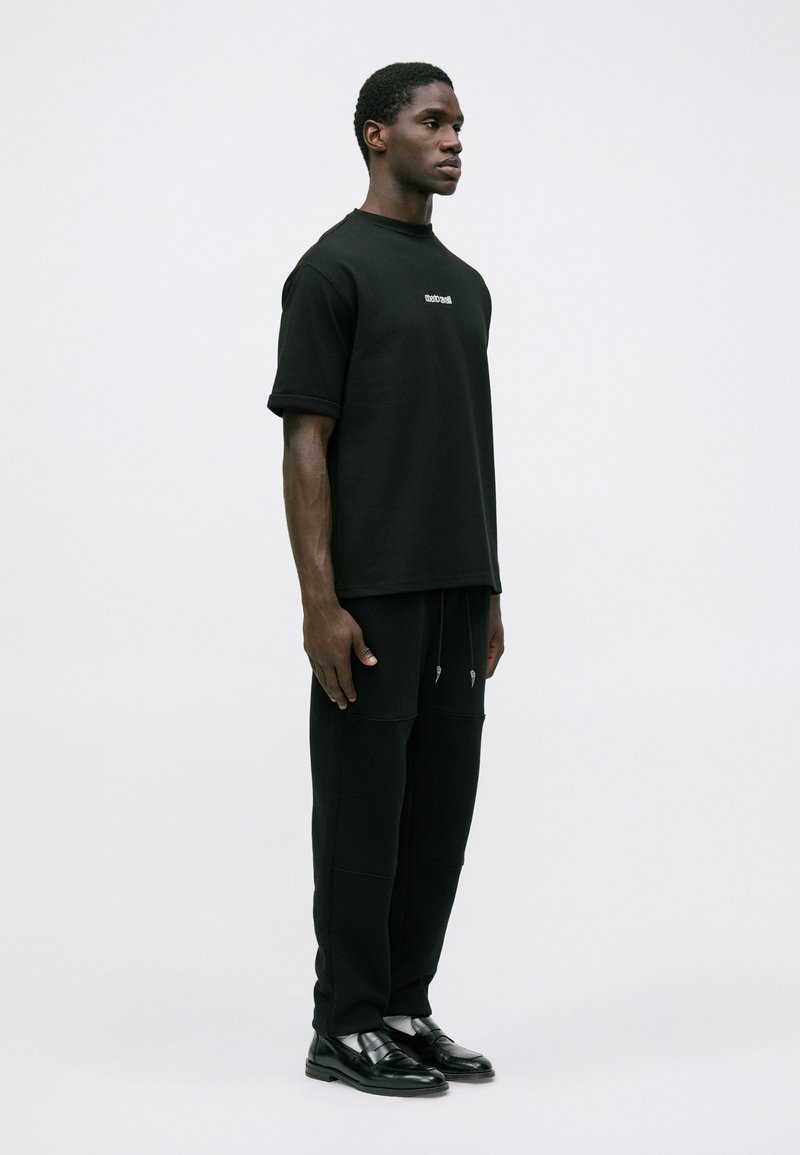 Homme se tenant de côté, portant une chemise noire à manches courtes, un pantalon noir avec cordons, des chaussettes blanches et des chaussures habillées noires sur un fond uni.