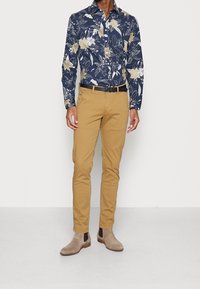 Camicia blu navy con motivo floreale e bottoni, pantaloni chino color sabbia aderenti, cintura nera e stivali Chelsea beige. Tessuto liscio, vestibilità su misura e texture sottili.