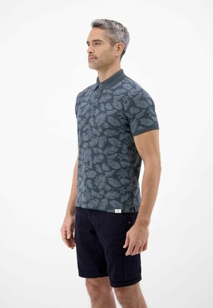 Mann steht seitlich, trägt ein dunkelblaues Polohemd mit floralem Muster und schwarze Cargo-Shorts vor einem einfarbigen weißen Hintergrund.