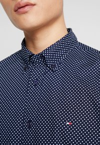 Camisa azul marino con lunares blancos, cuello con botones y dos botones en la parte frontal, con un pequeño logo en rojo y blanco.