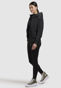 Veste à capuche noire avec poches avant et détails zippés, associée à des leggings noirs et des baskets montantes vert olive avec semelles blanches.