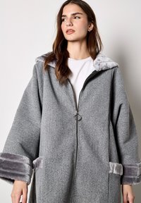Cappotto in misto lana grigio con cappuccio e polsini foderati in pelliccia sintetica argento, dotato di cerniera frontale e design a vestibilità ampia.
