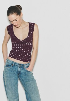 Woman with dark hair in a bun wearing a purple polka-dot sleeveless top and loose blue jeans, standing against a plain white background.