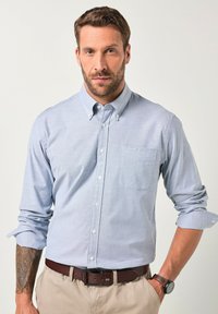 Camisa de botón azul claro con un bolsillo en el pecho, mangas largas remangadas en los puños, complementada con un cinturón marrón y un reloj.