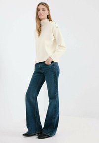 Pulover crem cu guler tip helancă și detalii cu nasturi, asortat cu blugi evazați de culoare albastru închis. Textură netedă, croială stilată și culori contrastante.