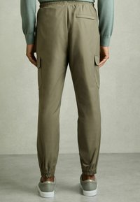 Pantaloni cargo verde măsliniu, cu talie elastică, gambe ajustabile și buzunare laterale, confecționați dintr-un material neted. Se văd adidași în partea de jos.
