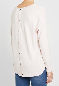 Jasnoróżowy sweter z zaokrąglonym tyłem, ozdobiony guzikami wzdłuż kręgosłupa. Miękka tekstura i luźny krój z długimi rękawami.