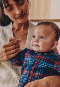 Dziecko w niebiesko-czerwonej koszuli w kratę z guzikami, o okrągłej twarzy i krótkich włosach, trzymane przez osobę w jasnym swetrze z grubym splotem.