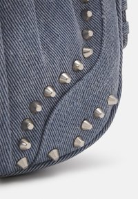 Denim tas met zilveren metalen studs langs de naad, met een gestructureerde blauwe stof die een ribbelpatroon creëert. Compact ontwerp en ronde vorm.