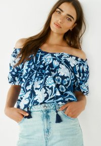 Blusa blu floreale off-the-shoulder con maniche a palloncino, tessuto texturizzato, frange decorative e una vestibilità comoda, abbinata a pantaloni shorts in denim di un blu chiaro.