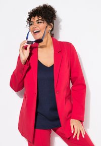 Felrood op maat gemaakte blazer met een enkele knoop, gedragen over een marineblauw geribbeld topje. Het model houdt een zonnebril vast en laat een krullend kapsel zien.