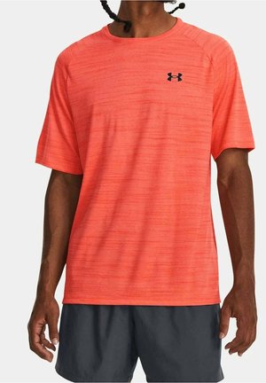 Mand iført en skarpt orange kortærmet sportstrøje med sort Under Armour-logo og mørkegrå shorts, stående mod en ensfarvet baggrund.