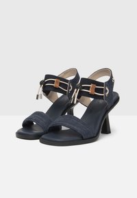 Mørkeblå hælede sandaler med stofremme i denim, en solid fodseng og dekorative hardware-detaljer på ankelremmen.