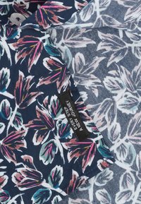 Bloem-shirt met marineblauwe stof en grote, kleurrijke bladeren in roze, groen en wit. Inclusief een zwart label met tekst.