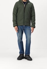 Mand iført en mørkegrøn hættejakke, blå jeans og sorte sneakers, der står op ad en hvid baggrund.