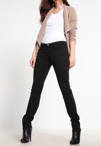 Jeans negros ajustados con una cintura de media altura, combinados con un cárdigan beige y una camiseta blanca. Llevados con botas negras de tacón. Textura suave.