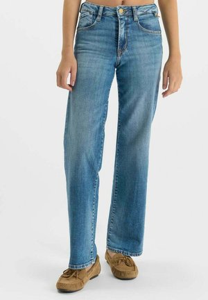 Vrouw draagt rechte blauwe spijkerbroek en zandkleurige suède mocassins, staand tegen een effen lichte achtergrond.