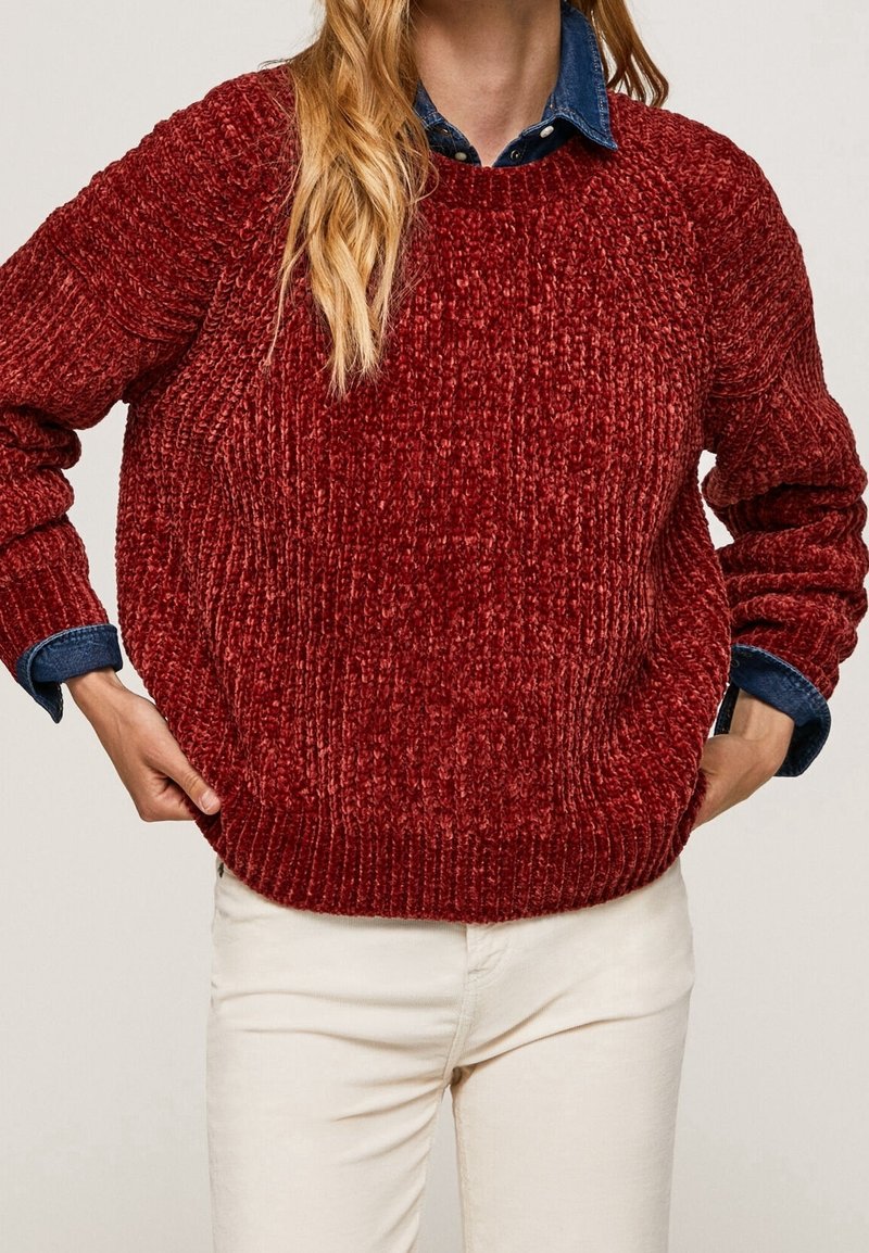 Personne portant un pull rouge en tricot par-dessus une chemise en jean bleu avec un pantalon de couleur claire, les mains dans les poches.