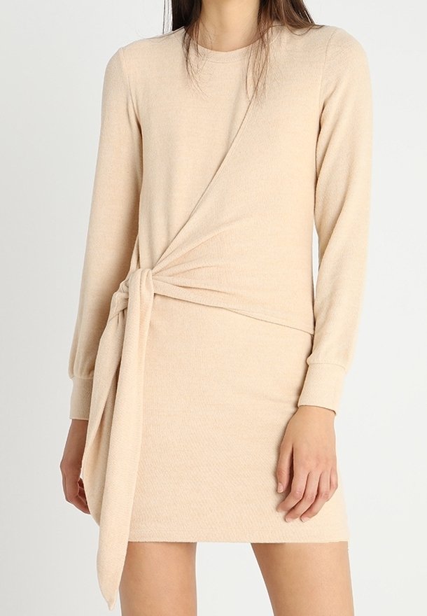 Femme portant une mini-robe beige à manches longues avec un détail noué sur le côté à la taille, debout devant un fond uni clair.