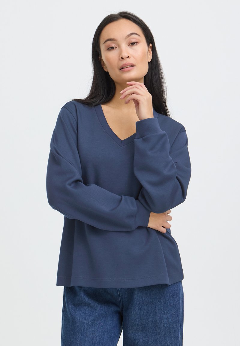 Femme aux longs cheveux foncés portant un haut bleu foncé à col en V à manches longues et un jean bleu, posant avec une main près du menton.