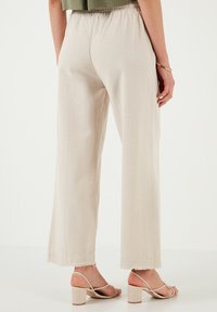 Beige weite Beine Hose mit strukturierter Oberfläche, ausgestattet mit einem ausgefransten Saum und einem elastischen Bund. Getragen mit sandalen mit Blockabsatz und Riemchen.