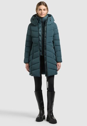 Femme portant une longue doudoune matelassée bleu sarcelle avec un col montant, des leggings noirs et des bottes noires montantes, debout sur un fond uni.