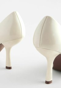 Chaussures à talons hauts beige avec une finition brillante, un bout pointu et des talons fins et effilés. Présente un accent marron à la base. Texture lisse.
