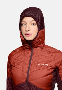 Giacca isolante da donna con una combinazione di colori maroon e ruggine, design trapuntato, cappuccio aderente e chiusura frontale con zip.