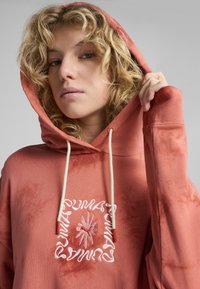 Kapuzen sweatshirt v rustovej farbe vyrobený z mäkkej tkaniny, s predným logom vyšitým v ružovej farbe a krémovými šnúrkami. Hladká textúra s uvoľneným strihom.
