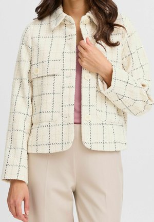 Frau trägt eine cremefarbene karierte, zumachenbare Jacke über einem malvenfarbenen Oberteil und beigen Hosen, hält die Jacke mit der rechten Hand am Kragen.