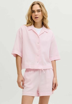 Giovane donna con lunghi capelli biondi che indossa una camicia rosa pastello ampia con bottoni e pantaloncini abbinati su uno sfondo semplice.