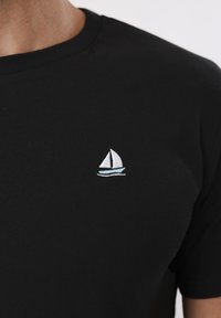 Μαύρο μπλουζάκι με μια μικρή λευκή και μπλε ιστιοφόρο κεντημένη στην αριστερή περιοχή του στήθους.