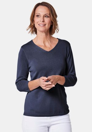 Maglione lavorato a maglia blu navy con maniche a tre quarti, scollatura a V con cuciture decorative e orlo aderente. Indossato sopra pantaloni bianchi.