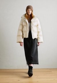 Veste en cream matelassée avec un col haut, associée à une jupe midi en denim noir. Un bonnet en tricot marron et des chaussures noires épaisses complètent le look.