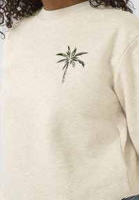 Sudadera beige hecha de una tela suave, con un diseño de palmera verde oscuro en el pecho. Cuello acanalado y corte relajado.