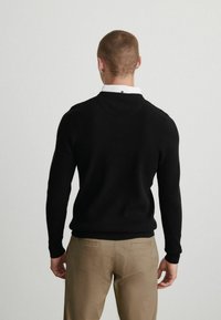 Sort strikket sweater med rund halsudskæring, der har ribbede mansjetter og kant. Båret over en hvid skjorte med krave, parret med khaki bukser.
