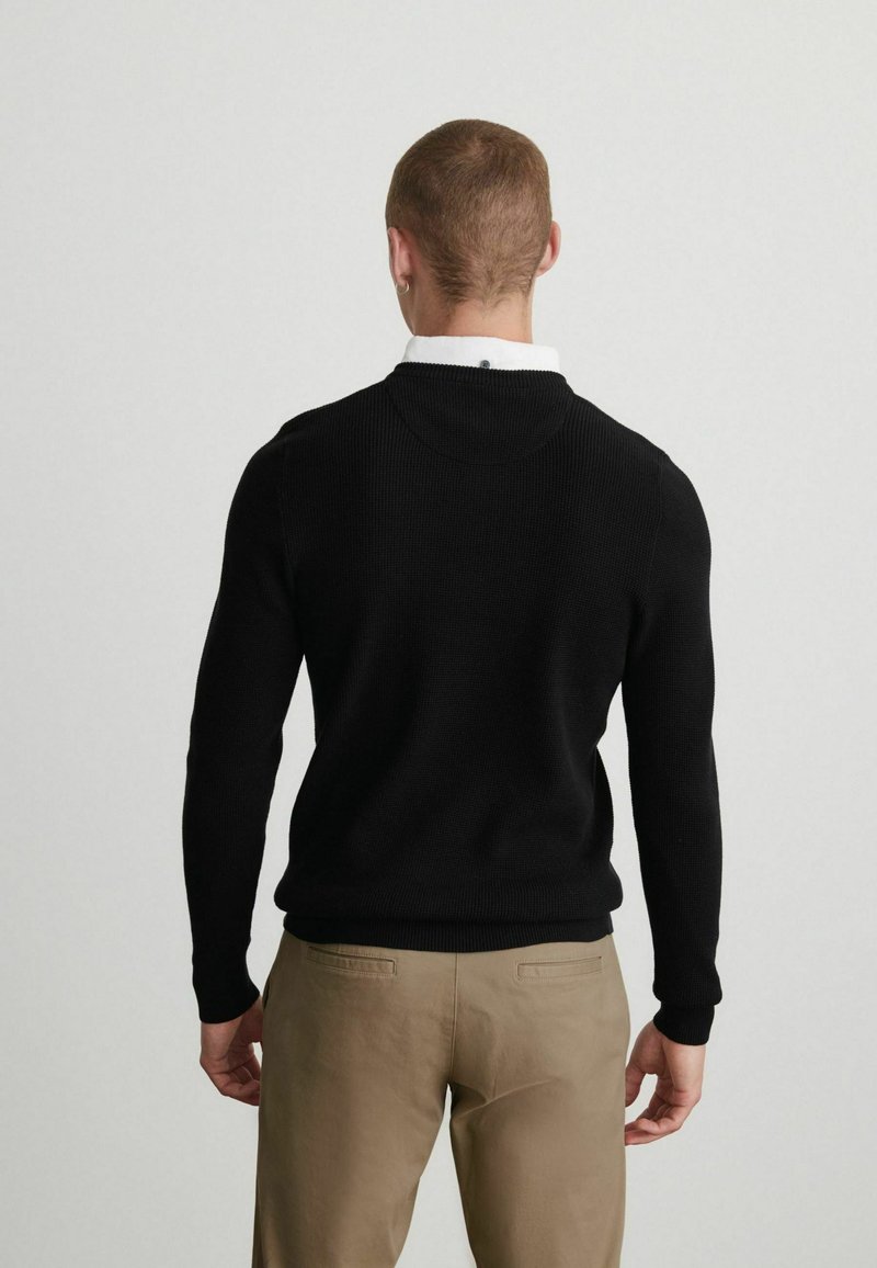 Sort strikket sweater med rund halsudskæring, der har ribbede mansjetter og kant. Båret over en hvid skjorte med krave, parret med khaki bukser.