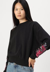 Μαύρη μπλούζα με μακριά μανίκια, διακοσμημένη με πολύχρωμη κεντημένη λουλούδια στο αριστερό μανίκι. Διαθέτει ραμμένο τελειώμα και μανίκια με ρίγες, φτιαγμένη από μαλακό ύφασμα.