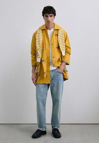 Chaqueta de algodón amarilla con cierres de toggles, superpuesta a una camisa blanca, combinada con vaqueros de denim claros y zapatos negros, y con una bufanda rayada.